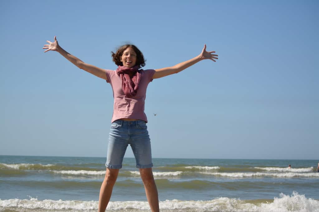 Eine Frau am Strand reißt die Arme hoch. Sie steht am Strand, dahinter ist das Meer.