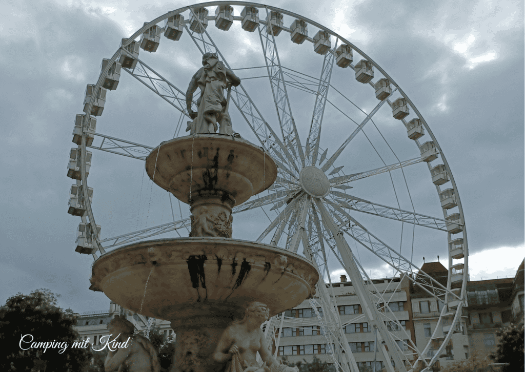Ein Springbrunnen und dahinter befindet sich ein Riesenrad
