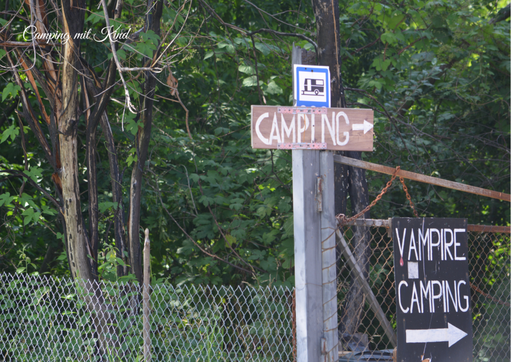 Vor einem Zaun befindet sich ein Holzpfahl. Darauf befindet sich ein Campingzeichen mit einem Pfeil. Dahinter sind Bäume.