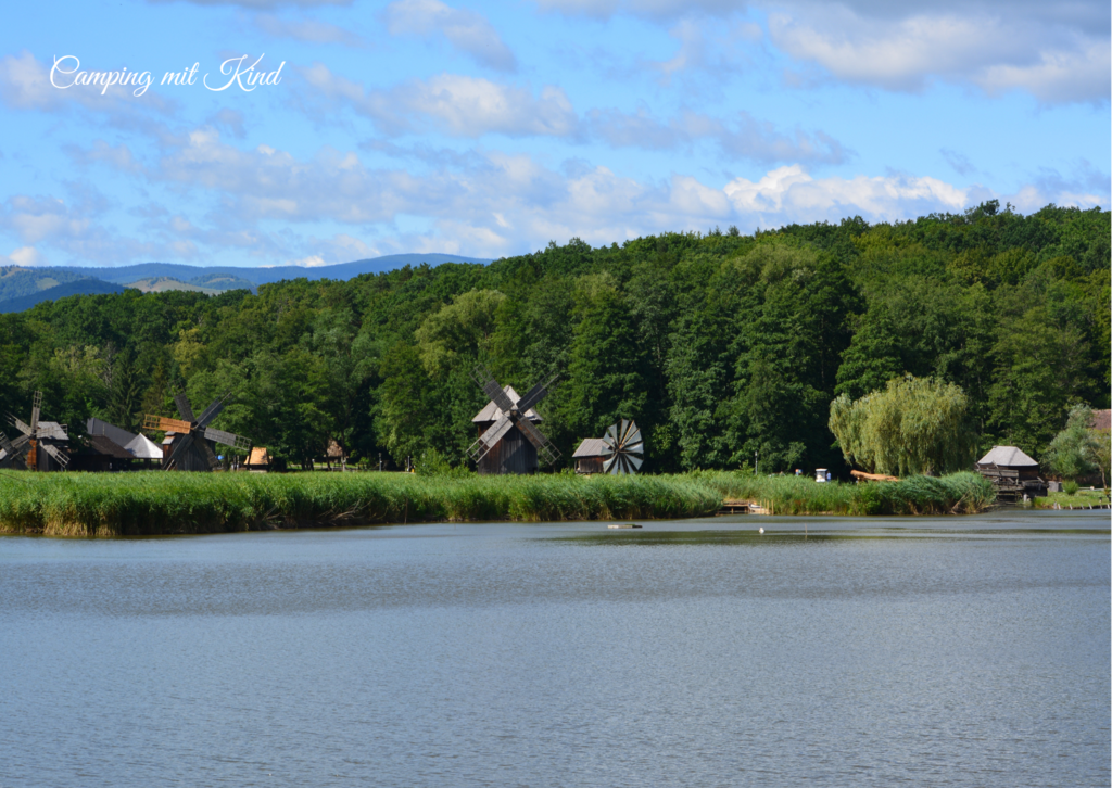 Im Vordergrund ist ein See. Dahinter befindet sich Windmühlen auf einer Wiese, dahinter grüne Wälder.