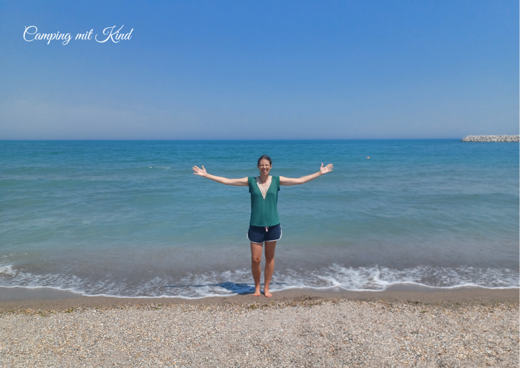 Eine Frau in kurzer Hose und T-Shirt steht am Strand und streckt ihre Arme auf die Seite.