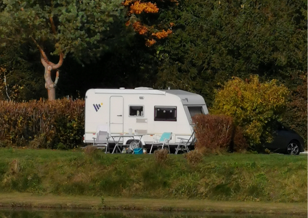 Ein Wohnwagen steht am See. Davor befinden sich Campingstühle und ein Tisch