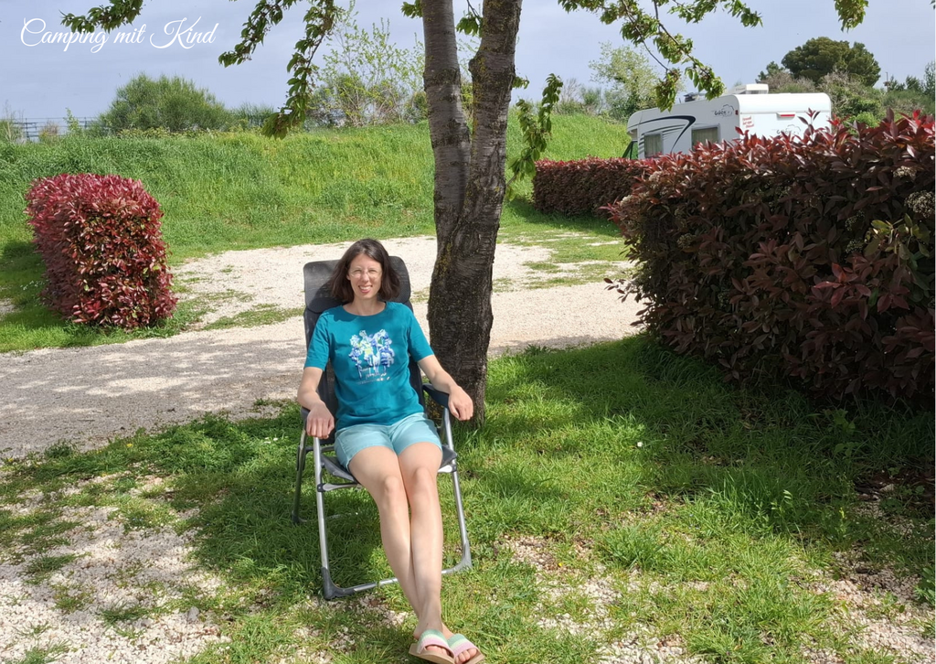 Monika sitzt auf einem Campingsessel neben einem Baum und einer Hecke. Sie trägt eine kurze Hose und ein T-Shirt.