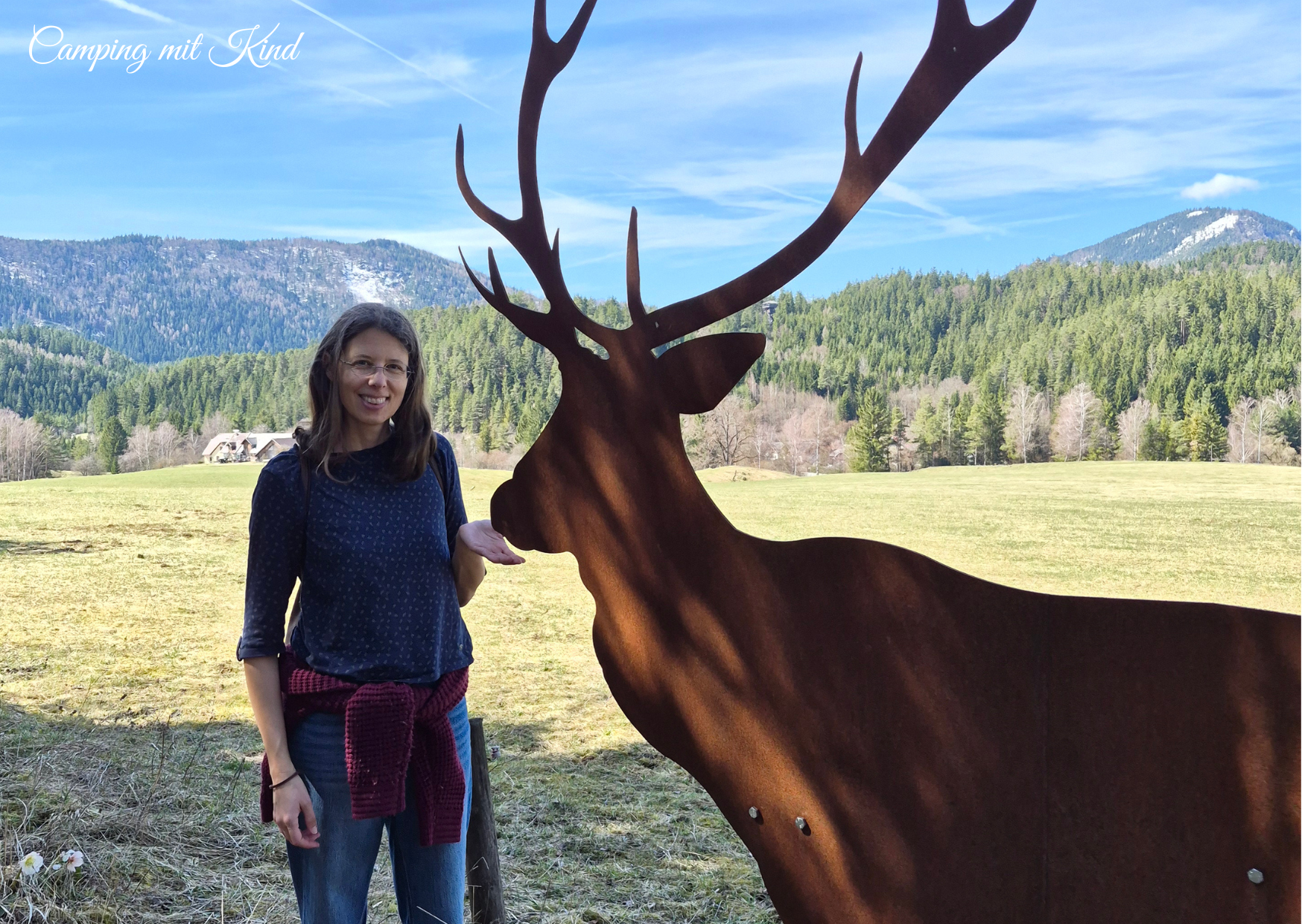 Monika steht auf einer Wiese, neben ihr steht ein Hirsch aus Metall, dahinter befindet sich ein schönes Panorama.