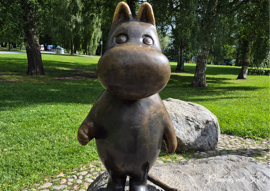 Eine Muminskulptur steht in einem Park. Dahinter sind Bäume, die Schatten machen