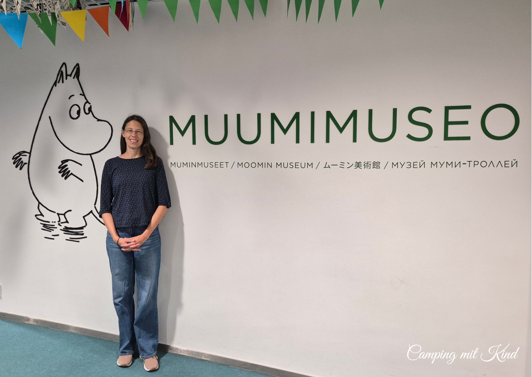 Moni steht an einer weißen Wand. Darauf steht Muumimuseo und ein Mumin ist abgebildet